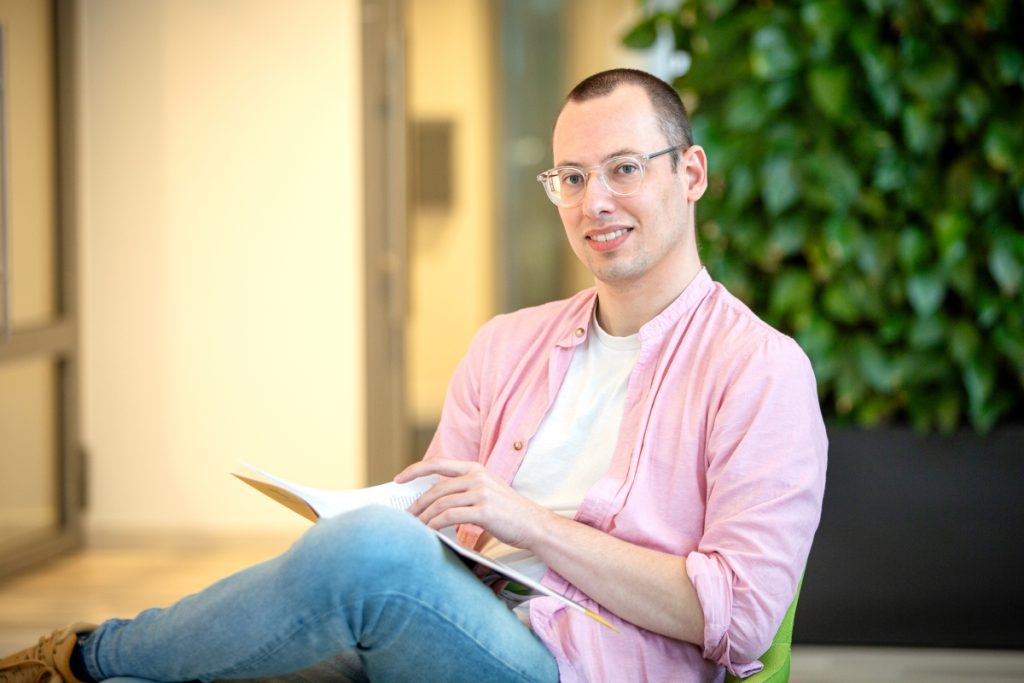 Een software engineer met een roze overhemd en bril zit op een groene stoel in de centrale hal bij Technolution, portret 3 van 5